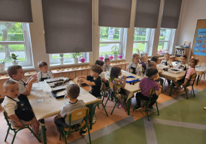 Dzieci siedzą przy stolikach i słuchają instrukcji.