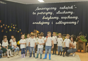 Dzieci stoją, śpiewają piosenkę i grają na instrumentach.