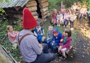 Dzieci siedzą na drewnianych pieńkach i słuchają skrzata