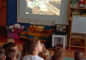 Dzieci siedzą na dywanie i oglądają prezentację
